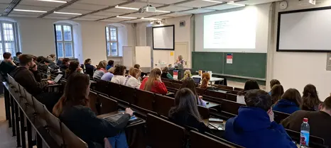 Das Foto zeigt den gro?en H?rsaal im Erdgeschoss des Geb?udes "An der Universit?t 5." Studierende sitzen auf stufig angeordneten Sitzb?nken und h?ren der Vortragenden, Cris Miller, eine wei?e Frau mit grauen, kinnlangen Haaren zu. Das Foto ist aus der rechten oberen Ecke des H?rsaals aufgenommen.